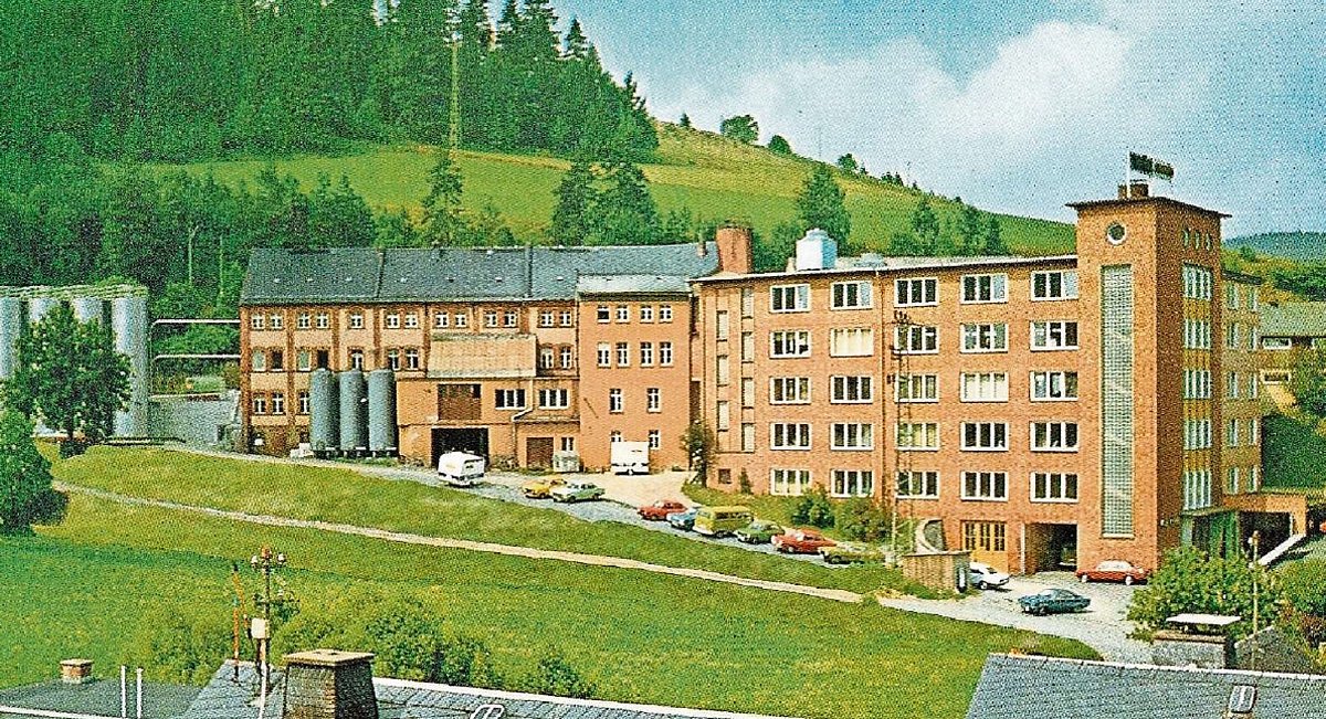 Ein großes Fabrikgebäude aus roten Ziegeln. Rundherum Wald, Wiesen und einzelne Autos. Das Foto ist aus dem Jahr 1985.