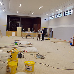 Zwei Männer mit einem Plan in den Händen stehen in einem großen Saal mit Bühne. Der Raum ist eine Baustelle. Es stehen und liegen Bauutensilien überall herum.