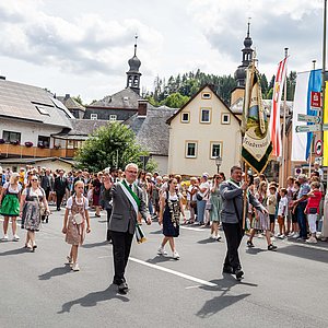 Zahlreiche Männer und Frauen in Schützenuniformen und Dirndln laufen auf der Straße einen Umzug. Sie alle lachen und winken den Menschen am Straßenrand zu.