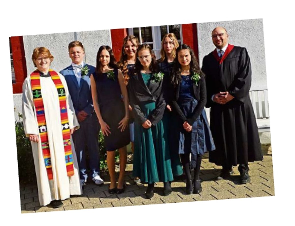Eine Gruppe Konfirmanden (5 junge Frauen und ein junger Mann) sowie eine Pfarrerin in weißem Talar und ein junger Mann in schwarzer Robe stehen vor einer Kirchenwand in Sonnschein und lächeln in die Kamera.
