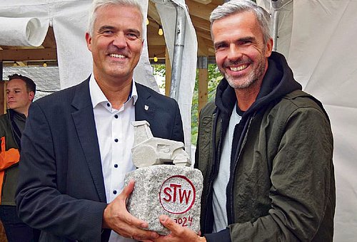 Zwei Männer stehen nebeneinander und halten gemeinsam einen mit roter Schrift gravierten Granitstein.