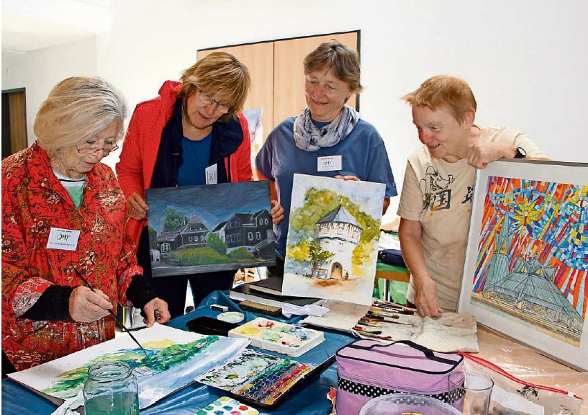 Vier Frauen stehen an einem Tisch voller gemalten Bilder. Drei von ihnen halten je ein Bild in den Händen. Sie schauen auf das Bild der vierten Frau, die etwas auf dem Bild zeigt. 