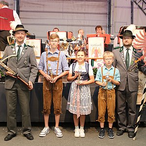 Vor einer Bühne in einem Zelt stehtn zwei Männer in Schützenuniform. Zwischen ihnen stehen zwei Jungen und ein Mädchen mit Pokalen.
